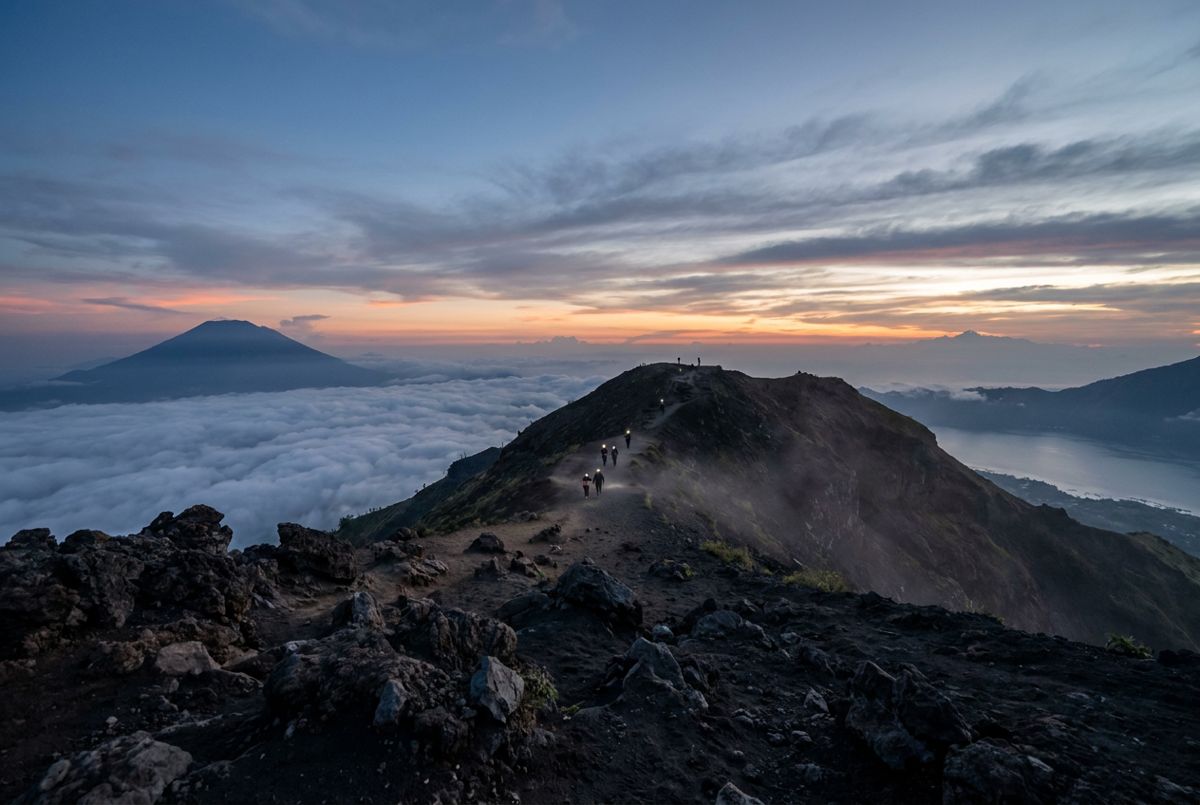 Sunrise over clouds and Lake Batur from the Mount Batur summit ridge, with distant hiker silhouettes