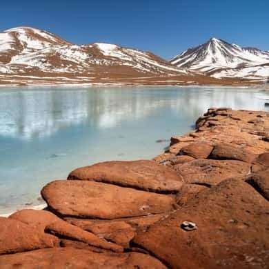 Red rocks and a lagoon beneath snow-capped volcanoes in the Atacama