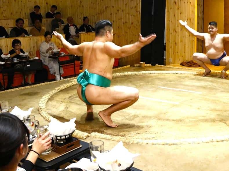 A sumo wrestler crouching in the ring while diners watch from tables