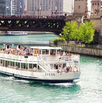 Chicago River cruise boat passing under DuSable Bridge with city buildings in the background.