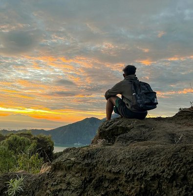 Person sitting on rocky ledge watching sunrise at Mount Batur, Bali.