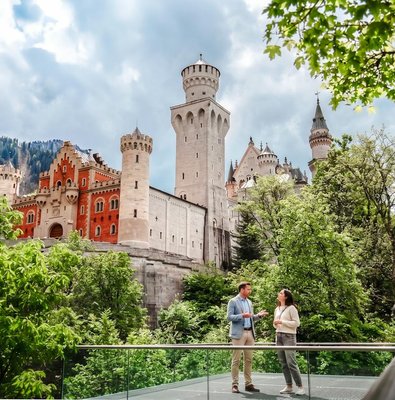 Tourists standing in front of Neuschwanstein Castle, Bavaria, surrounded by lush greenery.